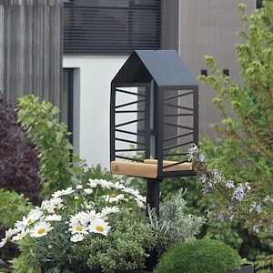 May include: A black metal bird feeder with a wooden perch, mounted on a black pole. The feeder has a house-like roof and a cage-like design. A black planter box with white daisies and green plants sits below.