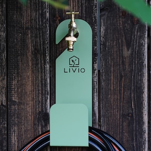 May include: A mint green metal hose holder with the word "LIVIO" printed on it, mounted on a weathered wooden fence. A brass faucet is attached to the holder, and a black and orange hose is coiled below.