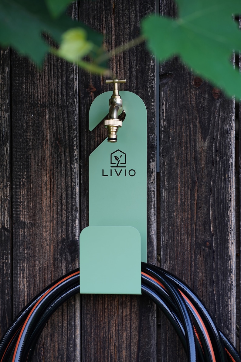 May include: A mint green metal hose holder with the word "LIVIO" printed on it, mounted on a weathered wooden fence. A brass faucet is attached to the holder, and a black and orange hose is coiled below.