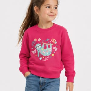 May include: A bright pink long-sleeve sweatshirt featuring a central graphic of a light blue sloth hanging from a branch, surrounded by colourful flowers, leaves, and butterflies arranged in a heart shape, paired with blue denim jeans.