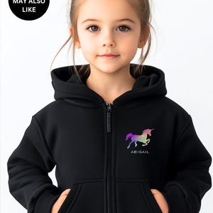 May include: A black zip-up hoodie featuring a hood, long sleeves, and two front pockets. On the left chest, there is a graphic of a unicorn in a rainbow gradient color scheme with the name ABIGAIL printed in white text underneath the unicorn.