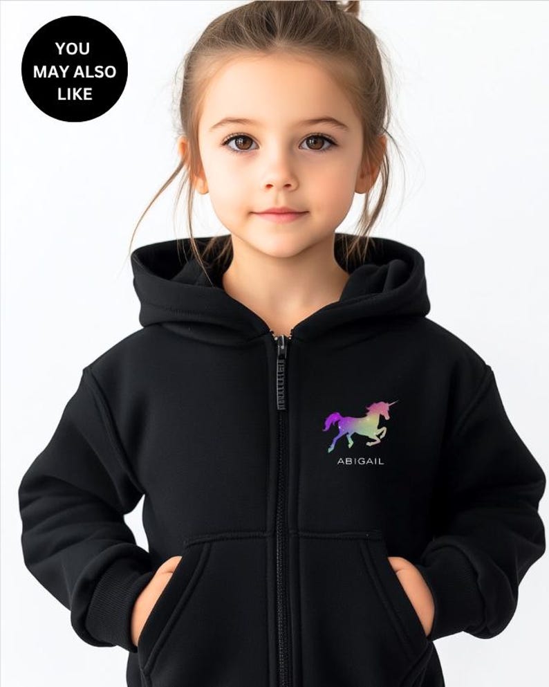 May include: A black zip-up hoodie featuring a hood, long sleeves, and two front pockets. On the left chest, there is a graphic of a unicorn in a rainbow gradient color scheme with the name ABIGAIL printed in white text underneath the unicorn.