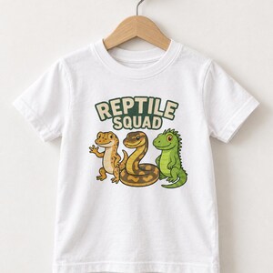Könnte beinhalten: Ein weißes kurzärmeliges T-Shirt mit einem Grafikdruck von drei Cartoon-Reptilien und dem Text REPTILE SQUAD in grünen Buchstaben. Die Illustration zeigt einen beigen Gecko, eine braun gemusterte Schlange und eine grüne Eidechse.