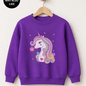 May include: A vibrant purple long-sleeve sweatshirt featuring a graphic print of a white unicorn with a multicolored mane, a pink flower, and small star accents. The sweatshirt has a crew neckline and ribbed cuffs and hem, hanging on a hanger.