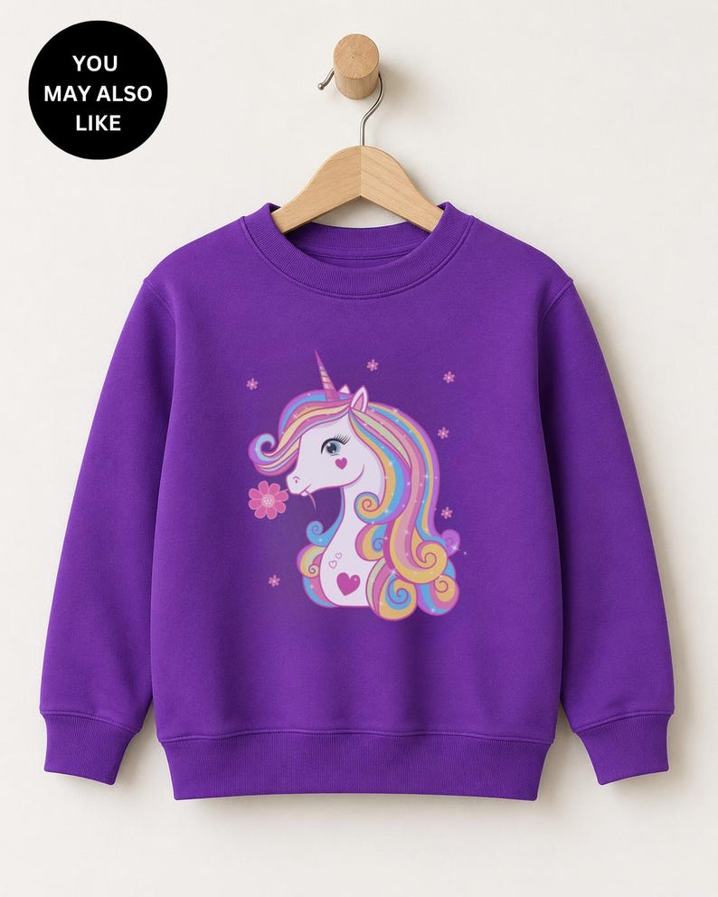 May include: A vibrant purple long-sleeve sweatshirt featuring a graphic print of a white unicorn with a multicolored mane, a pink flower, and small star accents. The sweatshirt has a crew neckline and ribbed cuffs and hem, hanging on a hanger.