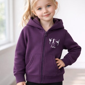 May include: A purple zip-up hoodie with a white ballet-themed graphic and the name "Isla" printed below. The hoodie has a hood, pockets, and a full-length zipper. The girl is wearing black leggings.