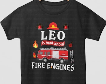 May include: A black short-sleeve t-shirt featuring a graphic print of a red fire engine, a firefighter helmet, flames, and a ladder. The text reads LEO is mad about FIRE ENGINES in white and red lettering against the dark fabric background.