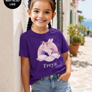 May include: A purple short-sleeved t-shirt featuring a graphic of a winged unicorn leaping over a rainbow and pink clouds. The name Freya is printed in white text below the graphic. The shirt is paired with light blue denim shorts.