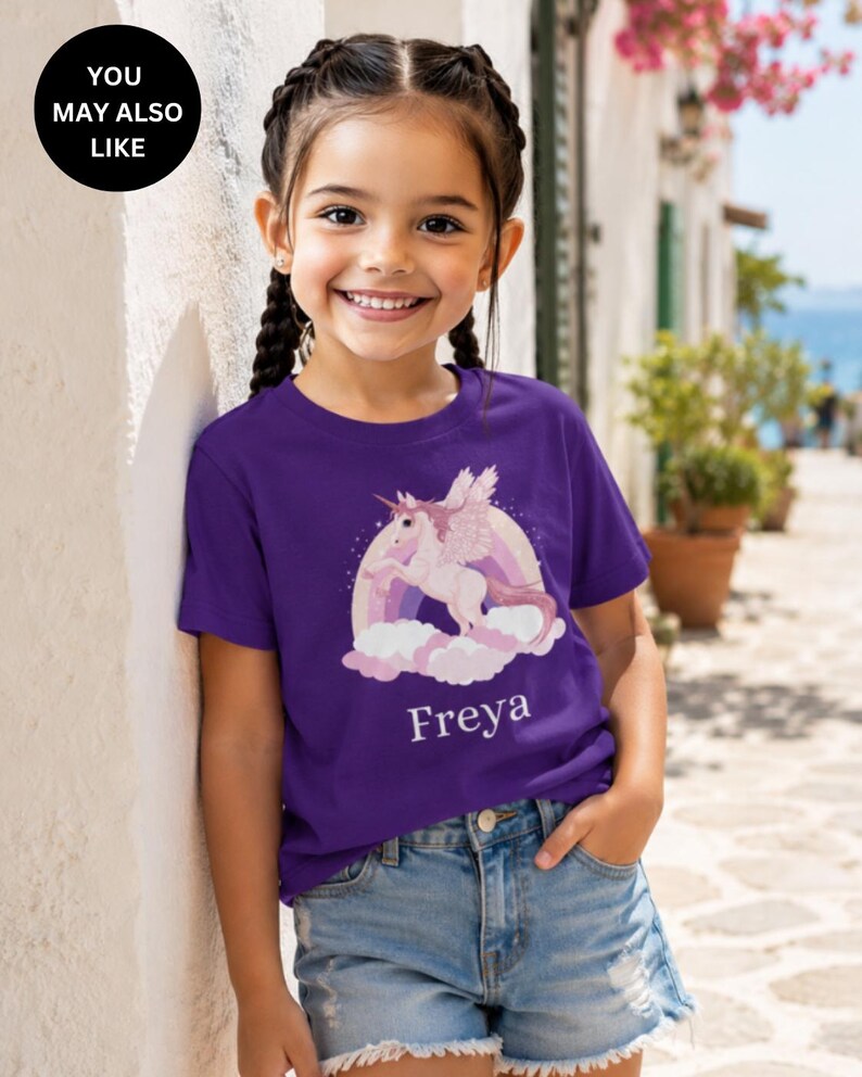 May include: A purple short-sleeved t-shirt featuring a graphic of a winged unicorn leaping over a rainbow and pink clouds. The name Freya is printed in white text below the graphic. The shirt is paired with light blue denim shorts.