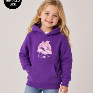 May include: A purple hooded sweatshirt featuring a graphic of a winged unicorn in front of a rainbow and clouds. The name Elodie is printed in white text below the graphic. The hoodie has long sleeves, a front kangaroo pocket, and ribbed cuffs.