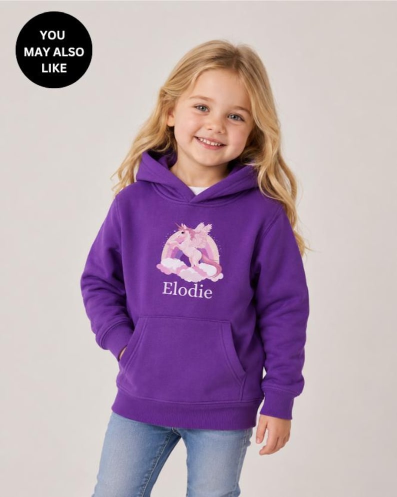May include: A purple hooded sweatshirt featuring a graphic of a winged unicorn in front of a rainbow and clouds. The name Elodie is printed in white text below the graphic. The hoodie has long sleeves, a front kangaroo pocket, and ribbed cuffs.
