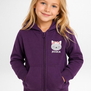 May include: A deep purple zip-up hoodie featuring a hood, long sleeves, and ribbed cuffs. On the left chest, there is a graphic of a gray cat face wearing a floral crown, with the name JESSICA printed in white capital letters underneath the cat.