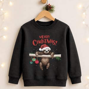 May include: A black long-sleeve sweatshirt featuring a graphic of a sloth wearing a red Santa hat while hanging from a branch. The text MERRY CHRISTMAS is printed in red above the sloth, which is surrounded by small white snowflakes and hanging ornaments.