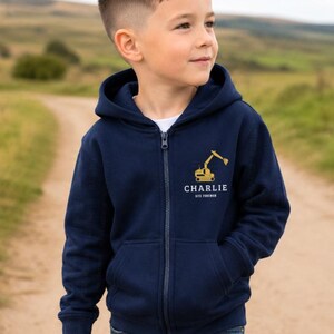 Könnte beinhalten: Marineblaue Kapuzenjacke mit Reißverschluss, goldener Bagger-Grafik und dem Namen "Charlie" mit "Site Foreman" darunter. Die Jacke hat eine Kapuze und Taschen. Darunter wird ein weißes T-Shirt getragen.