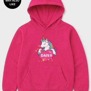 May include: A bright fuchsia long-sleeve hooded sweatshirt featuring a central graphic of a white unicorn with a rainbow mane and a gold horn. The unicorn rests on a large pink heart containing the white text DAISY, surrounded by small yellow stars.