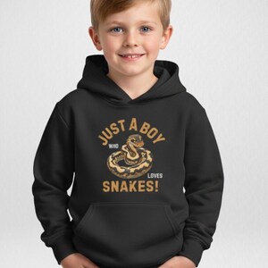 May include: Black hooded sweatshirt with the text "JUST A BOY WHO LOVES SNAKES!" in gold lettering. The graphic features a detailed illustration of a coiled snake in shades of brown and tan. The sweatshirt has a front pocket.