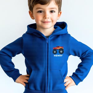 May include: A royal blue zip-up hoodie featuring a monster truck design on the chest. The monster truck is red and yellow with black wheels. The name "HENRY" is printed below the graphic in white.