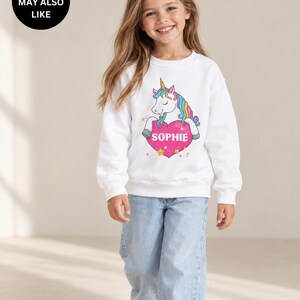 May include: A white long-sleeve sweatshirt featuring a graphic of a unicorn with a rainbow mane resting its front hooves on a large pink heart. The heart contains the name SOPHIE in white capital letters, surrounded by small yellow stars.