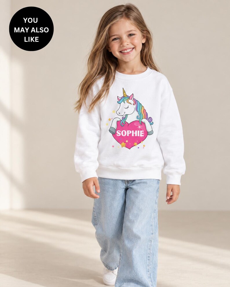 May include: A white long-sleeve sweatshirt featuring a graphic of a unicorn with a rainbow mane resting its front hooves on a large pink heart. The heart contains the name SOPHIE in white capital letters, surrounded by small yellow stars.
