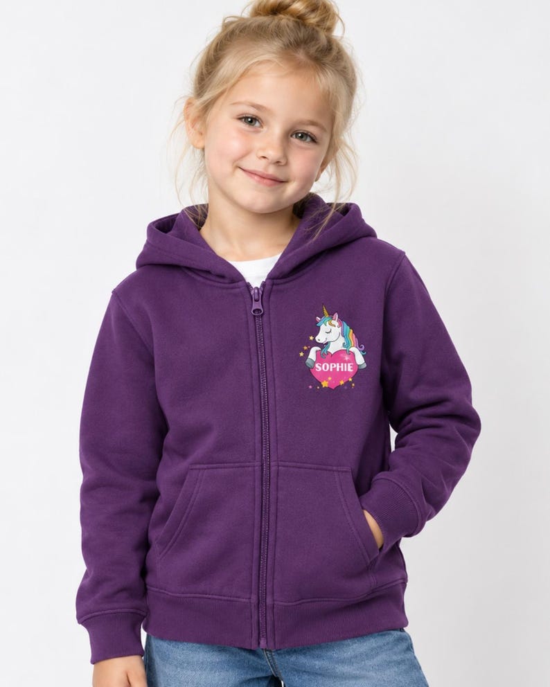 May include: A deep purple zip-up hoodie featuring a graphic on the left chest of a white unicorn with a rainbow mane, positioned above a pink heart containing the name SOPHIE in white capital letters, surrounded by small yellow stars.