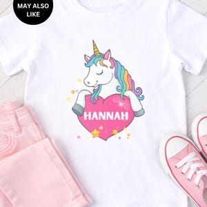 May include: A white short-sleeved t-shirt featuring a graphic of a unicorn with a rainbow mane and a gold horn. The unicorn is resting its front hooves on a large pink heart that has the name HANNAH printed in white capital letters.