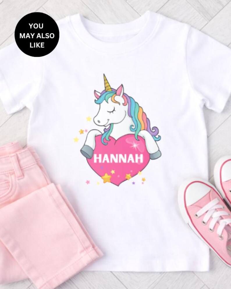 May include: A white short-sleeved t-shirt featuring a graphic of a unicorn with a rainbow mane and a gold horn. The unicorn is resting its front hooves on a large pink heart that has the name HANNAH printed in white capital letters.