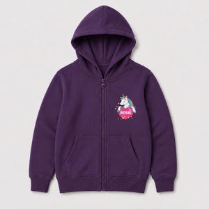 May include: A deep purple zip-up hoodie featuring a hood, long sleeves, and two front pockets. On the left chest, there is a graphic of a white unicorn with a colorful mane and a gold horn, positioned above a pink heart containing the name ROSIE.