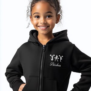 May include: A black zip-up hoodie featuring a white graphic of cheerleaders and the name "Aisha" in script. The hoodie has a front zipper, a hood, and two front pockets. The child is smiling.