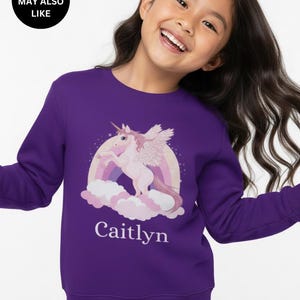 May include: A purple long-sleeve sweatshirt featuring a graphic print of a light pink winged unicorn standing on clouds in front of a rainbow. The name Caitlyn is printed in white text below the unicorn graphic on the front of the garment.