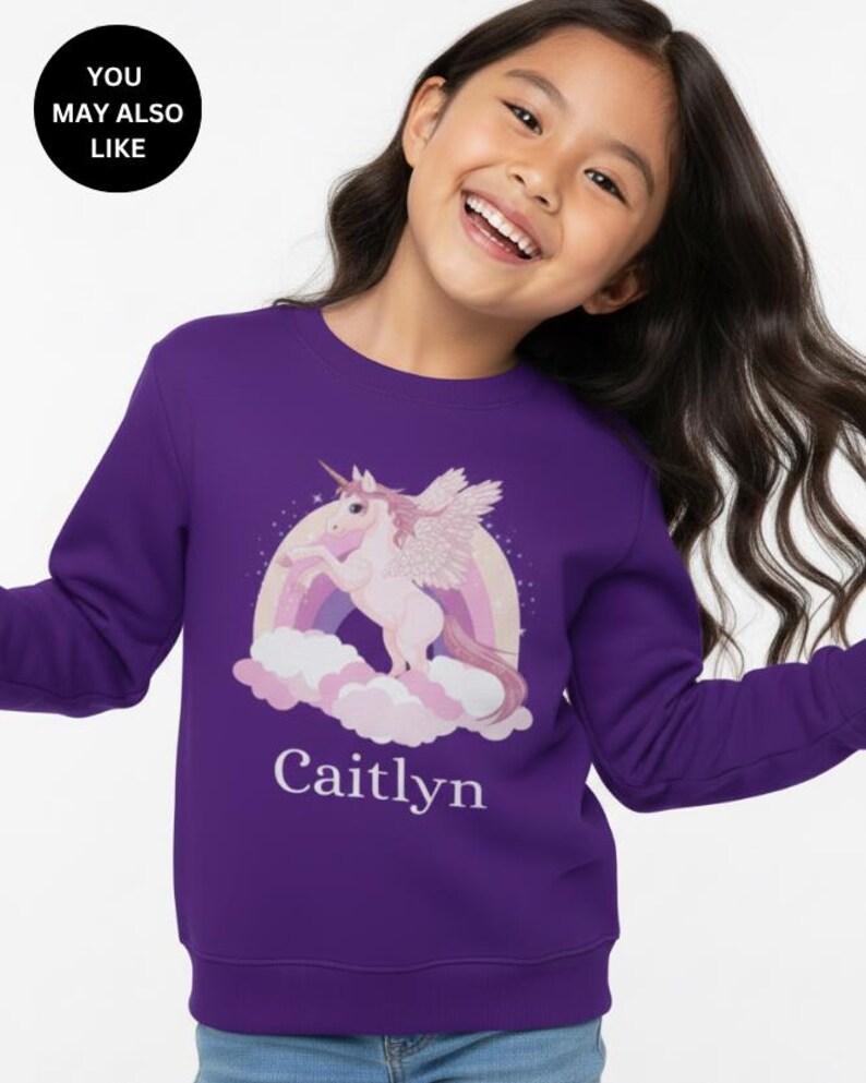 May include: A purple long-sleeve sweatshirt featuring a graphic print of a light pink winged unicorn standing on clouds in front of a rainbow. The name Caitlyn is printed in white text below the unicorn graphic on the front of the garment.