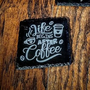 May include: Two black slate coasters with white text. One coaster says "Life Begins After Coffee" with a coffee cup graphic. The other coaster has a red and white graphic that says "Coffee". A green mug with a red and white "Coffee" graphic sits on the coaster.
