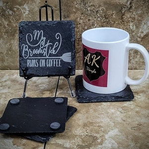 May include: A white ceramic mug with a black and red logo that says "AK Vinyls". The mug is sitting on a black slate coaster. A black slate coaster with the text "My Broomstick Runs on Coffee" is on a small easel.