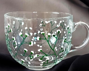 May include: Clear glass teacup with a handle, hand-painted with a delicate floral design. White flowers and green stems adorn the cup's exterior. The cup is approximately 4 inches in diameter.