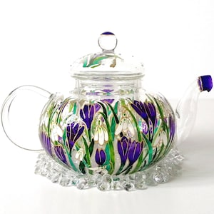 May include: A clear glass teapot with a floral design featuring purple crocuses and white snowdrops. The teapot has a glass lid and a glass base.