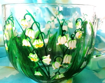May include: A clear glass mug with a hand-painted design of white lily of the valley flowers and green leaves. The mug has a handle and is sitting on a blue and white patterned fabric.