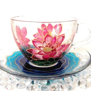 Puede incluir: Un juego de taza y platillo de té de vidrio transparente. La taza presenta un diseño de flor de loto rosa pintado a mano con detalles dorados. El platillo tiene un patrón de olas azules y doradas. El juego está sobre un lecho de piedras decorativas transparentes.