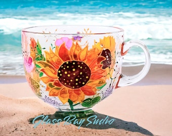 May include: A clear glass teacup with a hand-painted sunflower design. The sunflower is yellow and orange with a dark brown centre. Other floral elements in pink, purple, and green are also painted on the cup. The text "Glass Bay Studio" is at the bottom.