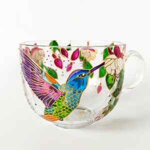 May include: A glass teacup with a hand-painted hummingbird design. The hummingbird is depicted in flight, with its wings spread and its beak extended towards a flower. The cup is decorated with pink, purple, green, and gold paint.