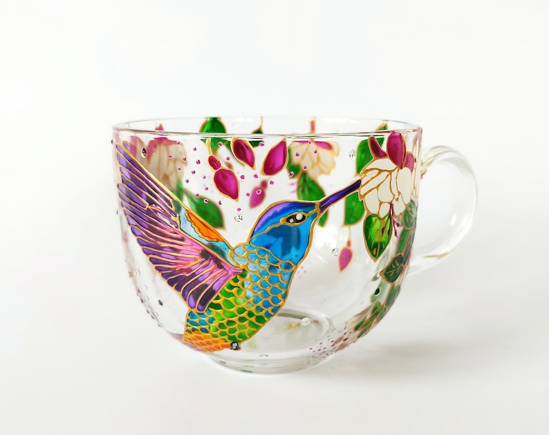 May include: A glass teacup with a hand-painted hummingbird design. The hummingbird is depicted in flight, with its wings spread and its beak extended towards a flower. The cup is decorated with pink, purple, green, and gold paint.