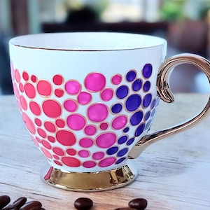 Könnte beinhalten: Weiße Keramik-Kaffeetasse mit goldfarbenem Rand und Henkel. Die Tasse ist mit einem Muster aus rosa, roten und violetten Kreisen verziert.