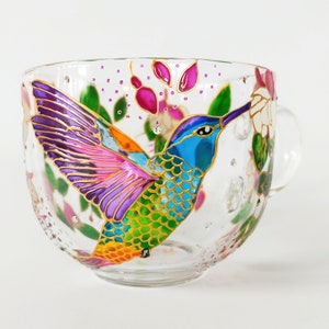 May include: A glass mug with a hand-painted hummingbird design. The hummingbird is depicted in vibrant colors, including purple, pink, green, blue, and orange. The mug has a handle and is decorated with flowers and leaves.