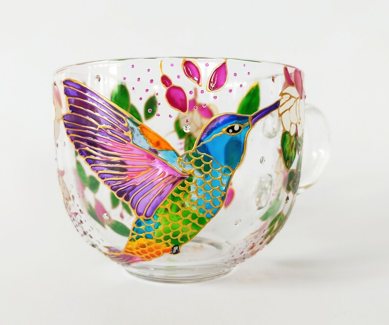 May include: A glass mug with a hand-painted hummingbird design. The hummingbird is depicted in vibrant colors, including purple, pink, green, blue, and orange. The mug has a handle and is decorated with flowers and leaves.