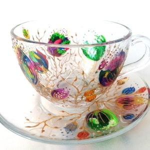 May include: A hand-painted glass teacup and saucer set with a floral design. The cup and saucer are decorated with gold branches and colorful flowers.