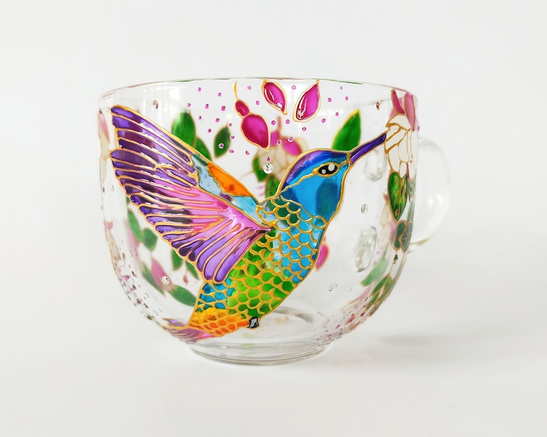 May include: A glass mug with a hand-painted hummingbird design. The hummingbird is depicted in flight with iridescent wings and a blue body. The mug is decorated with pink and green floral accents.