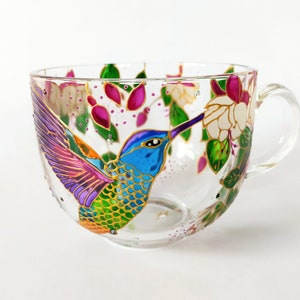May include: A glass mug with a hand-painted hummingbird design. The hummingbird is depicted in vibrant colors, including blue, green, purple, and orange, with gold accents. The hummingbird is shown with its beak in a pink flower.