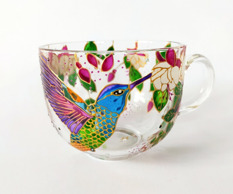 May include: A glass mug with a hand-painted hummingbird design. The hummingbird is depicted in vibrant colors, including blue, green, purple, and orange, with gold accents. The hummingbird is shown with its beak in a pink flower.