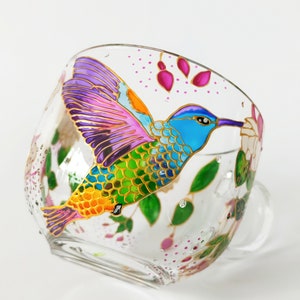 May include: A glass teacup with a hand-painted hummingbird design. The hummingbird is depicted in vibrant colors, including purple, blue, green, and yellow, with gold accents. The cup features a floral design with pink and green accents.