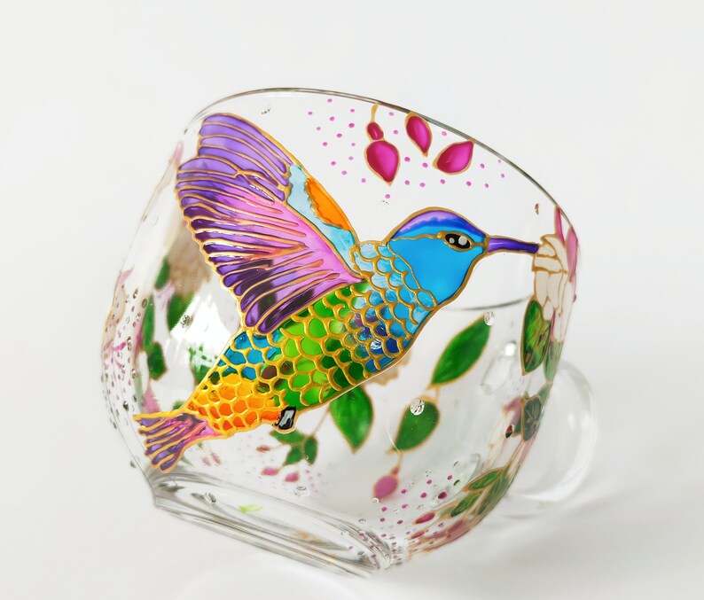 May include: A glass teacup with a hand-painted hummingbird design. The hummingbird is depicted in vibrant colors, including purple, blue, green, and yellow, with gold accents. The cup features a floral design with pink and green accents.