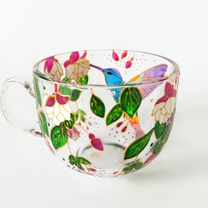 May include: A clear glass teacup with a painted design of a hummingbird in flight surrounded by pink and white flowers and green leaves.