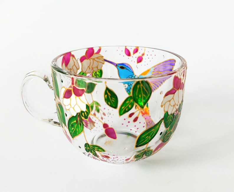 May include: A clear glass teacup with a painted design of a hummingbird in flight surrounded by pink and white flowers and green leaves.
